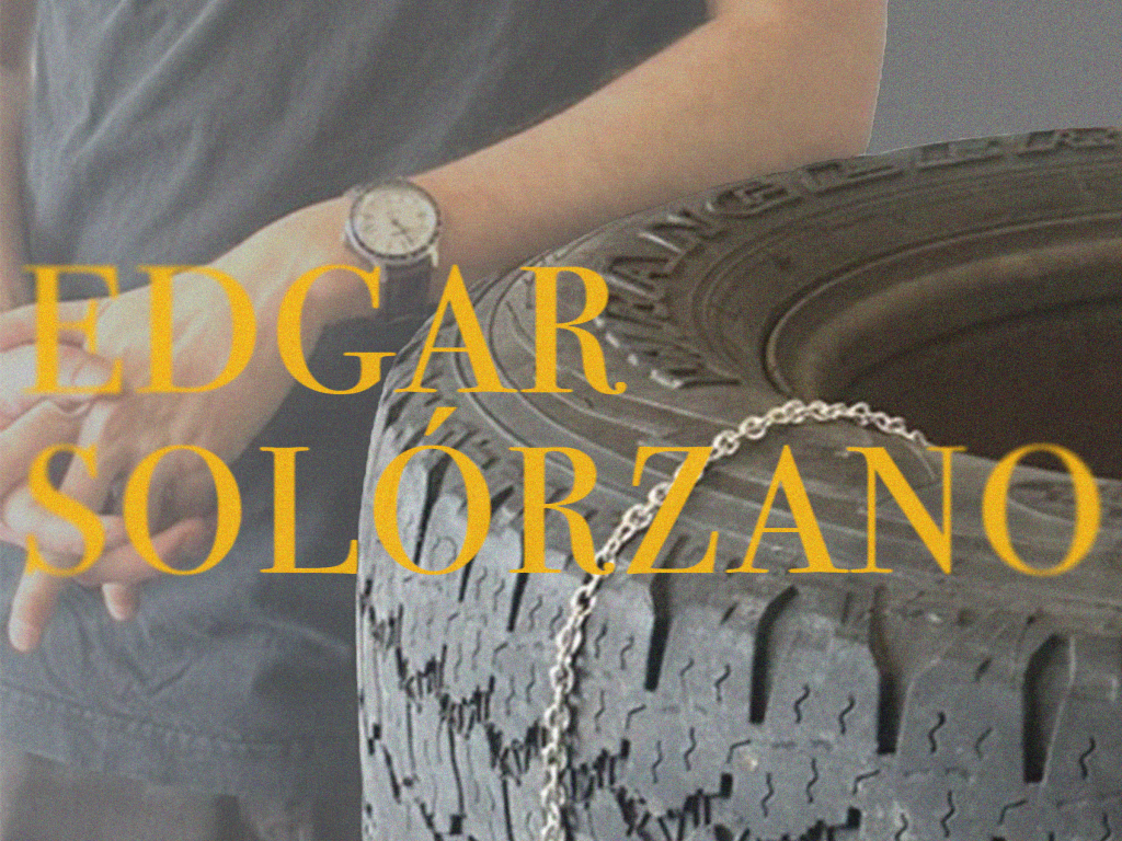 Image of the artist's torso and hands resting on a tire in his studio, with a thin silver chain laying across its surface. Over the image text reads "Edgar Solorzano"